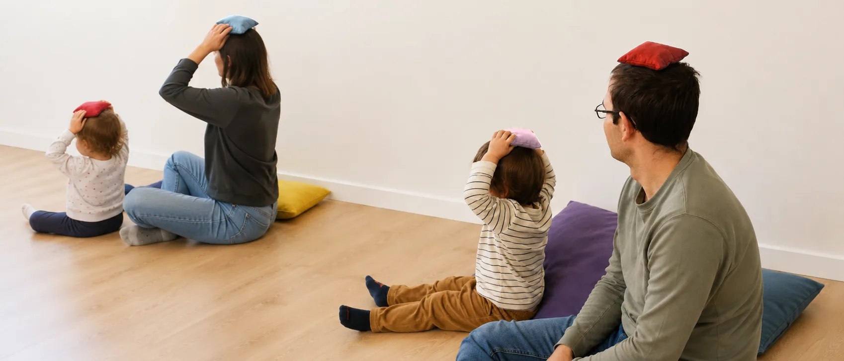 Dos adultos con un niño y una niña pequeños, sentados en el suelo de espaldas sobre cojines, cada uno con un saquito de tela en equilibrio sobre la cabeza